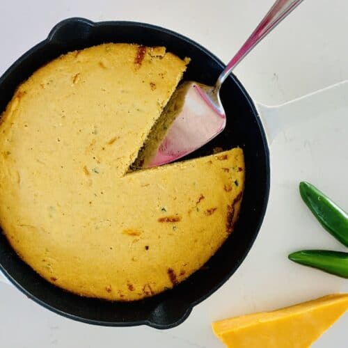 Birds Eye View: gluten-free jalapeño cheddar cornbread in a cast-iron skillet with white enamel. One slice is missing and a metal cake lifter is in its place. A wedge of cheddar and 2 green jalapeños are on the counter.