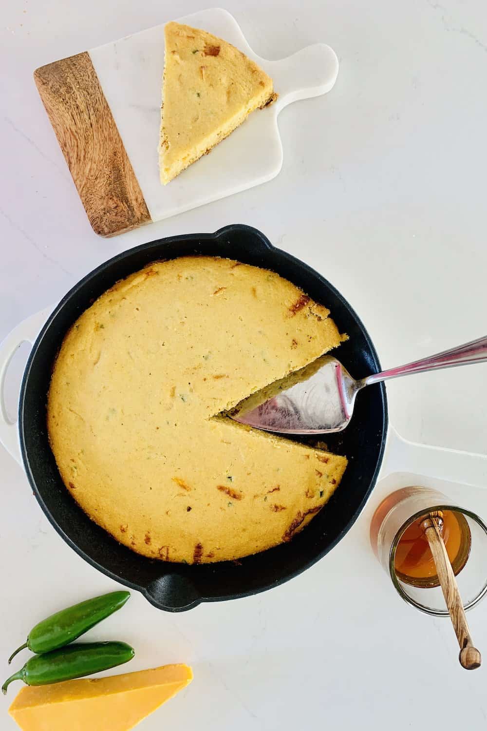 Birds Eye View: gluten-free jalapeño cheddar cornbread in a cast-iron skillet with white enamel. One slice is missing and a metal cake lifter is in its place. A slice of the cornbread on a wooed and marble cuttling board, a wedge of cheddar, 2 green jalapeños, and a glass of honey are on the counter.
