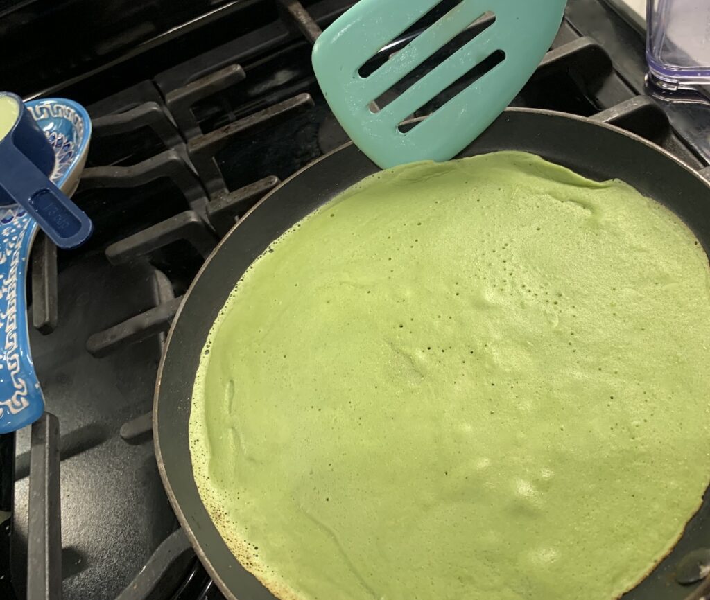 An aqua spatula lifting the edge of a bright green crepe from the crepe pan.