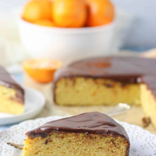 Slice of orange cake with chocolate ganache. Background includes the rest of the cake and a bowl of oranges.