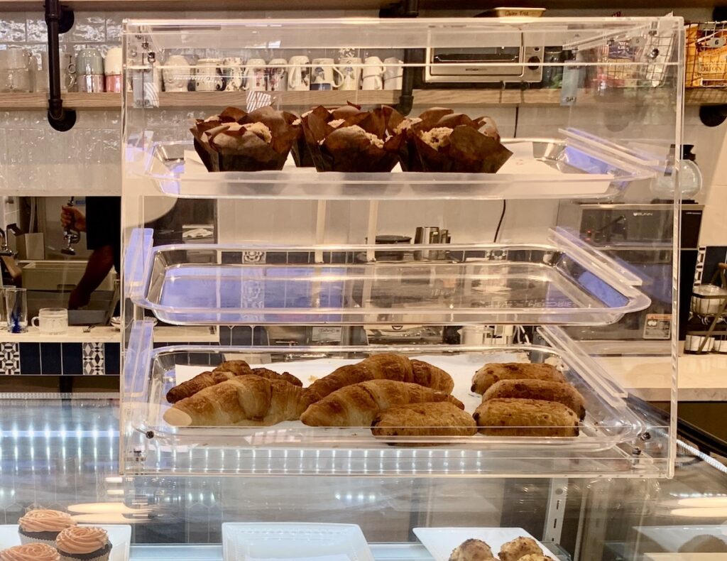 A plastic display case with 3 shelves. The top shelf has muffins. The middle shelf is empty. The bottom shelf has croissants and pain au chocolat.