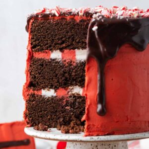 3-layeer chocolate peppermint cake with red frosting, red and white frosting between layers, crushed candy cane on top and chocolate ganache. A slice is removed revealing the inside layers.