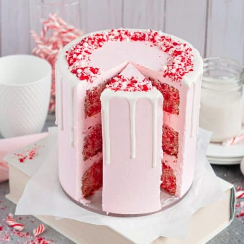 Candy cane cake with 3-red & white cake layers, pink frosting, crushed candy cane and white ganache rim. A slice has been cut and a smaller slice in its place allowing you to see the inside layers of the cake.