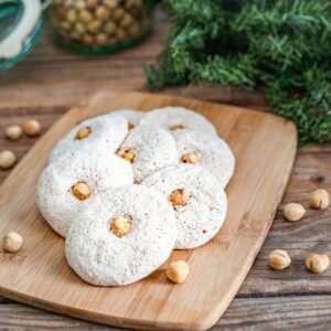 White hazelnut cookies with a hazelnut in the center of each cookie and scattered on the table and cutting board.