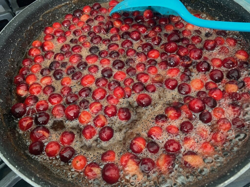 Cranberries cooking in bubbling, brown liquid in a frying pan, being stirred by an aqua spoon.