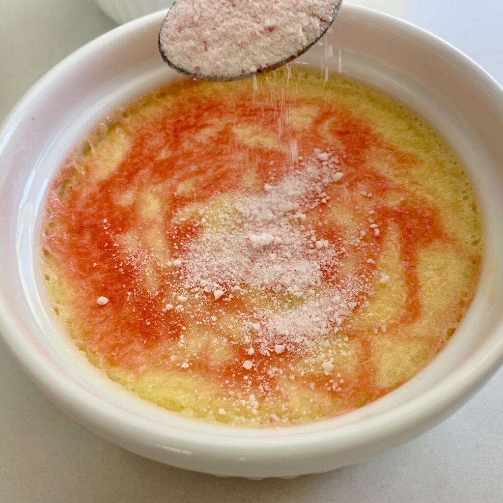 A spoon sprinkling crushed candy canes on top of swirled red and cream creme brûlée.