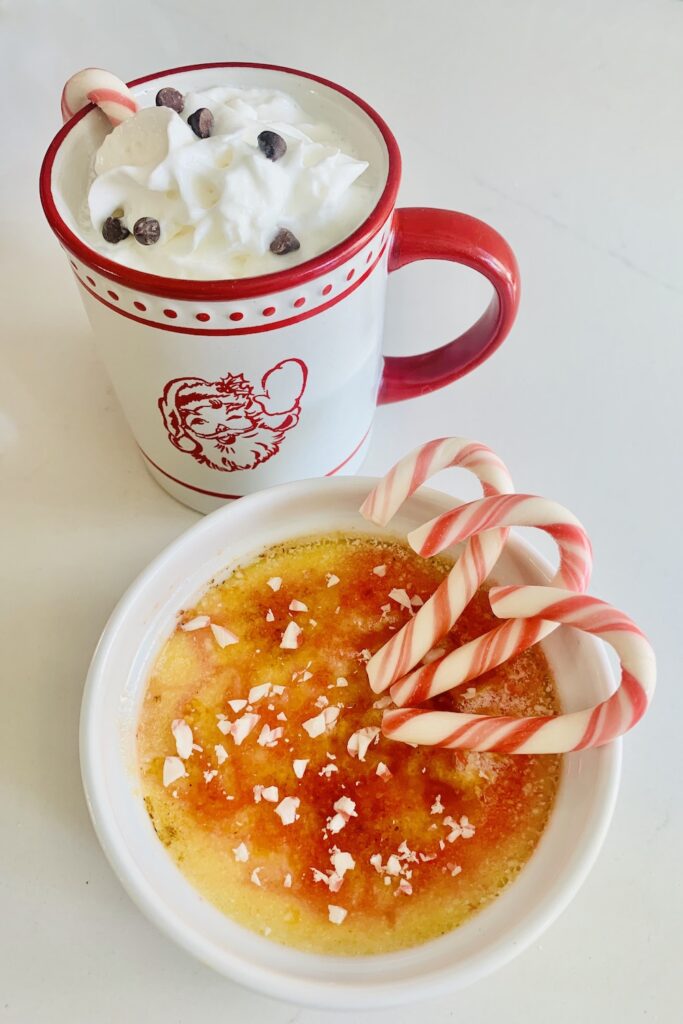 A cup of white hot chocolate in a Santa mug, topped with whipped cream, chocolate chips and a mini candy cane, next to candy cane creme brûlée topped with crushed candy canes and 3 mini candy canes.
