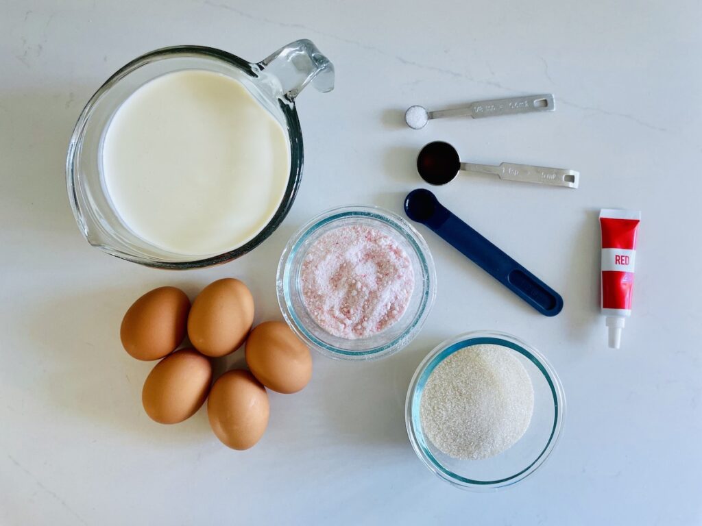 Birds eye view of ingredients on a counter: cream, 5 brown eggs, crushed candy canes, sugar, red food coloring tube, 3 measuring spoons with salt and liquids.