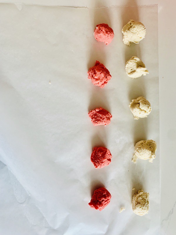 A row of white dough balls next to a row of red/pink dough balls, on parchment paper.