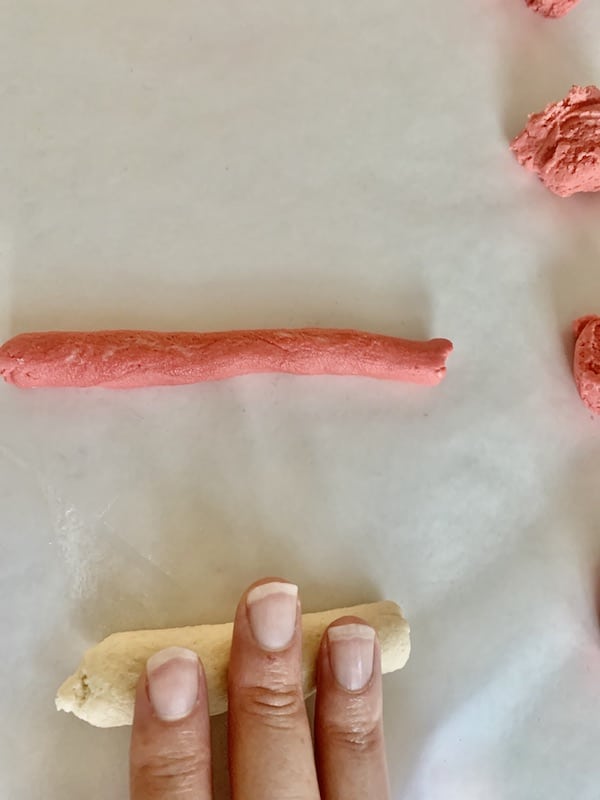 Overhead view: fingers rolling out white dough strip. Background, rolled out red dough strip and balls of red dough on the side.