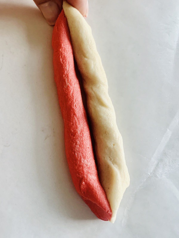 Overhead view: a strip of red dough next to white dough with the ends pressed together. Fingers pressing the top ends together.