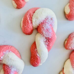 Bird's eye view: gluten-free candy cane cookies (white and red/pink dough twisted into a candy cane shape and covered with crushed candy cane sprinkles) scattered on a white counter.