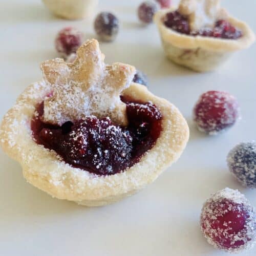 Gluten-Free Cranberry tart topped with a pie-crust snowflake cut-out. Sugared cranberries in the foreground and more cranberry tarts in the background.