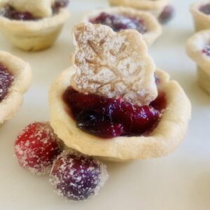 Gluten-Free Cranberry tart topped with a pie-crust leaf cut-out. Sugared cranberries in the foreground and more cranberry tarts in the background.