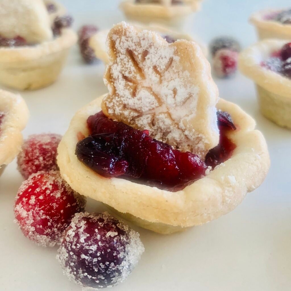 Gluten-Free Cranberry tart topped with a pie-crust leaf cut-out. Sugared cranberries in the foreground and more cranberry tarts in the background.