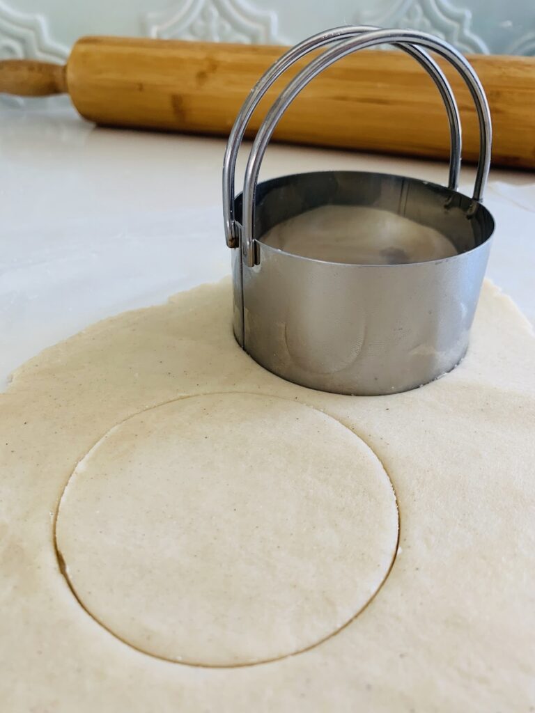Biscuit cutter on pie dough with one circle already cut. Rolling pin in the background.