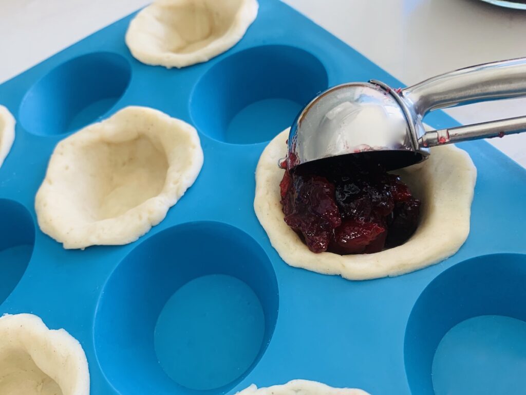 Bue mini silicone muffin pan with every other spots filled with pie crust and a cookie scooper filling one of the mini crusts with a scoop of cranberry sauce.