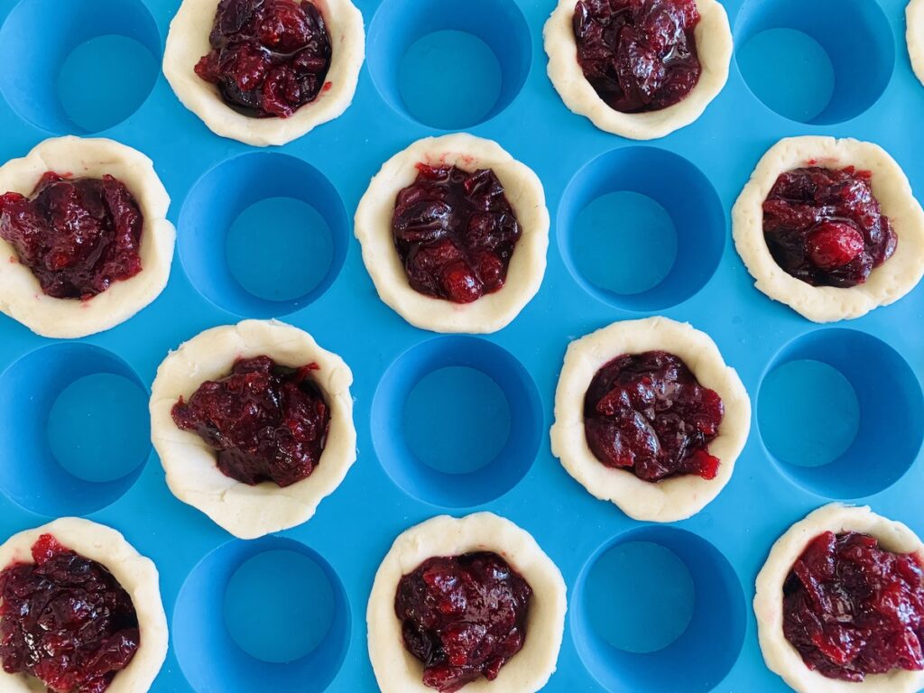 Birds eye view: blue mini silicone muffin pan with 12 of the spots filled with pie crust and cranberry sauce and 12 empty.