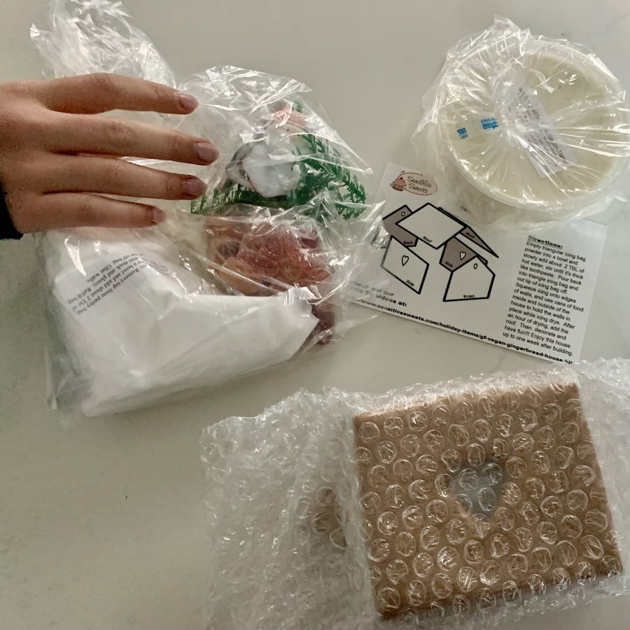Hand reaching for contents of gluten-free gingerbread house on the counter: piping bag of white powder for icing, bag of candy, bag of trees and santa/snowmen rings, card with instructions, container of frosting and bubble-wrapped house walls.