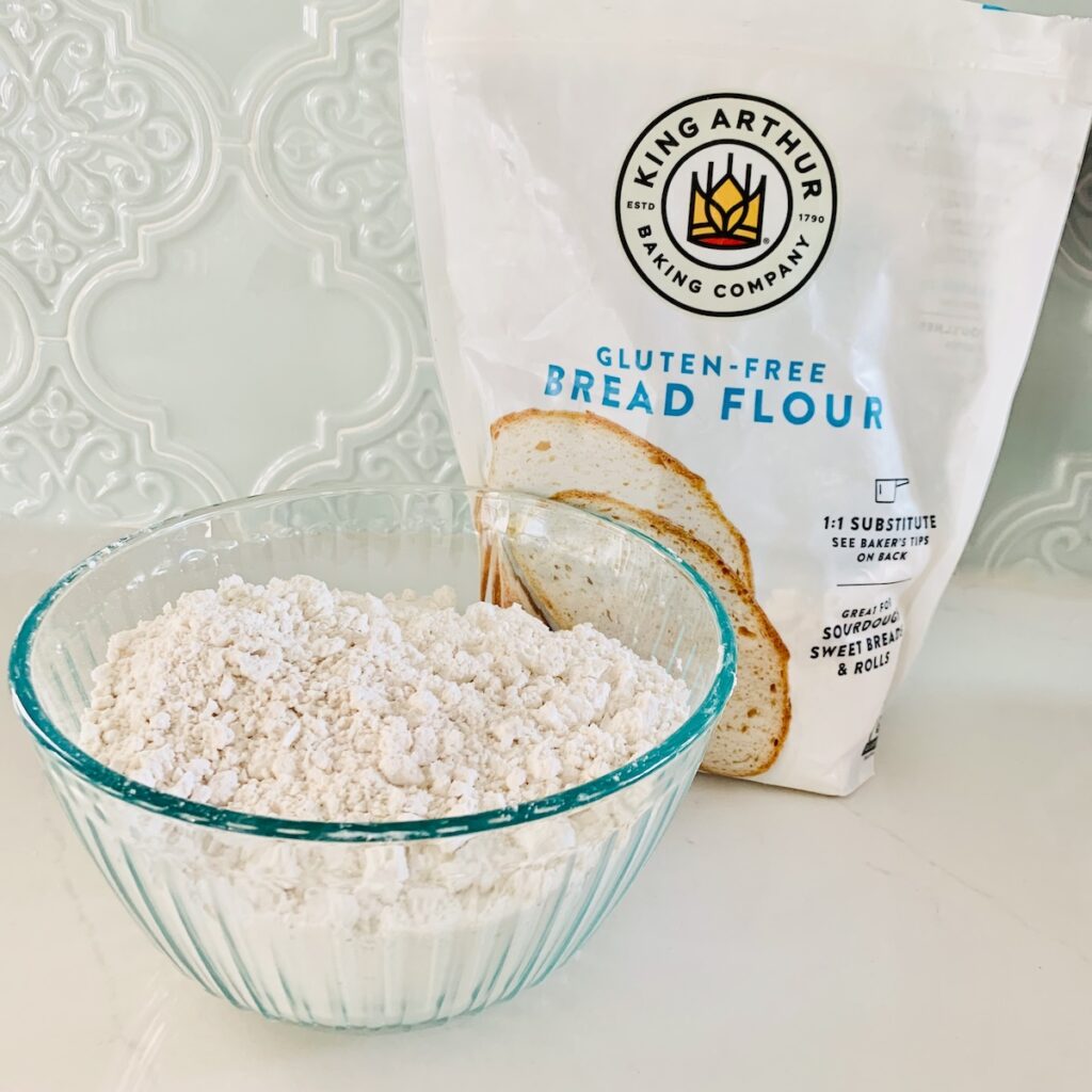 A glass bowl of flour in front of a bag of King Arthur Gluten-Free Bread Flour.