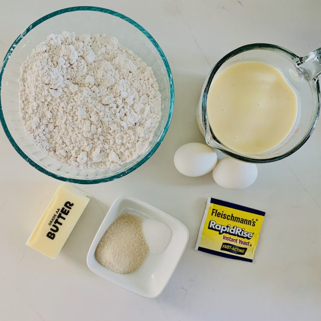 Birds Eye view of ingredients: flour, eggnog, 2 white eggs, pack of RapidRise Yeast, bowl with sugar and salt, and stick of butter.