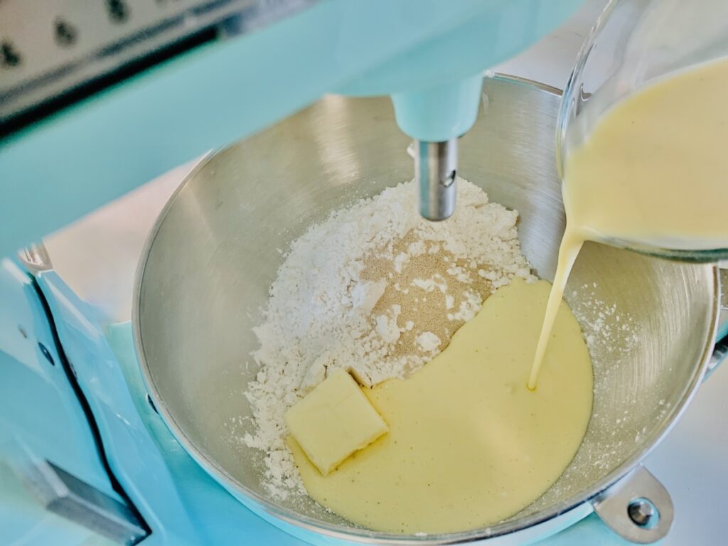 Ingredients (flour, yeast, butter) in a the mixing bowl of an aqua-colored mixer, with eggnog being poured into the bowl.
