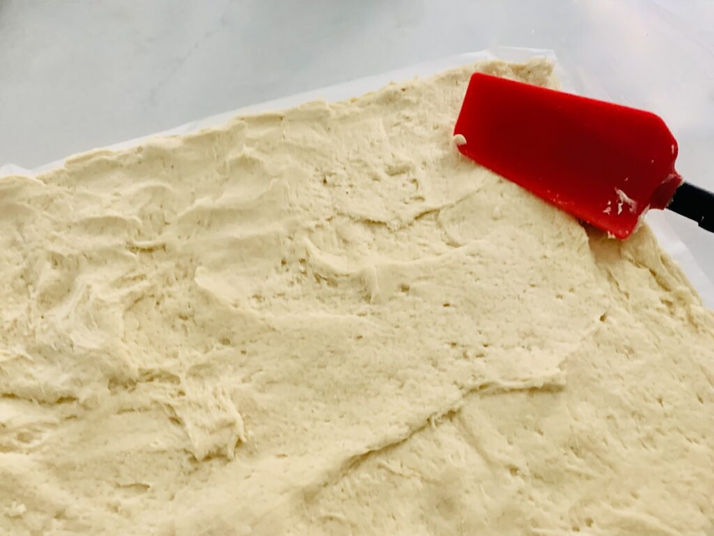 A red spatula spreading cream-colored dough into a rectangle-shape on parchment paper.