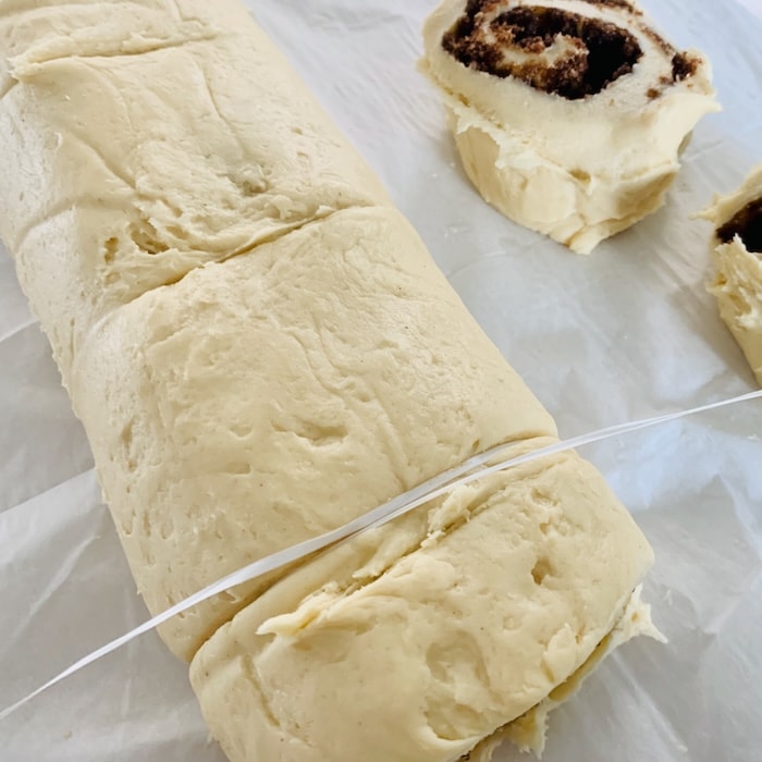 A cinnamon roll log with floss wrapped around the bottom one inch of the log and the ends being pulled to slice off a cinnamon roll. Part of two cut rolls in the background.