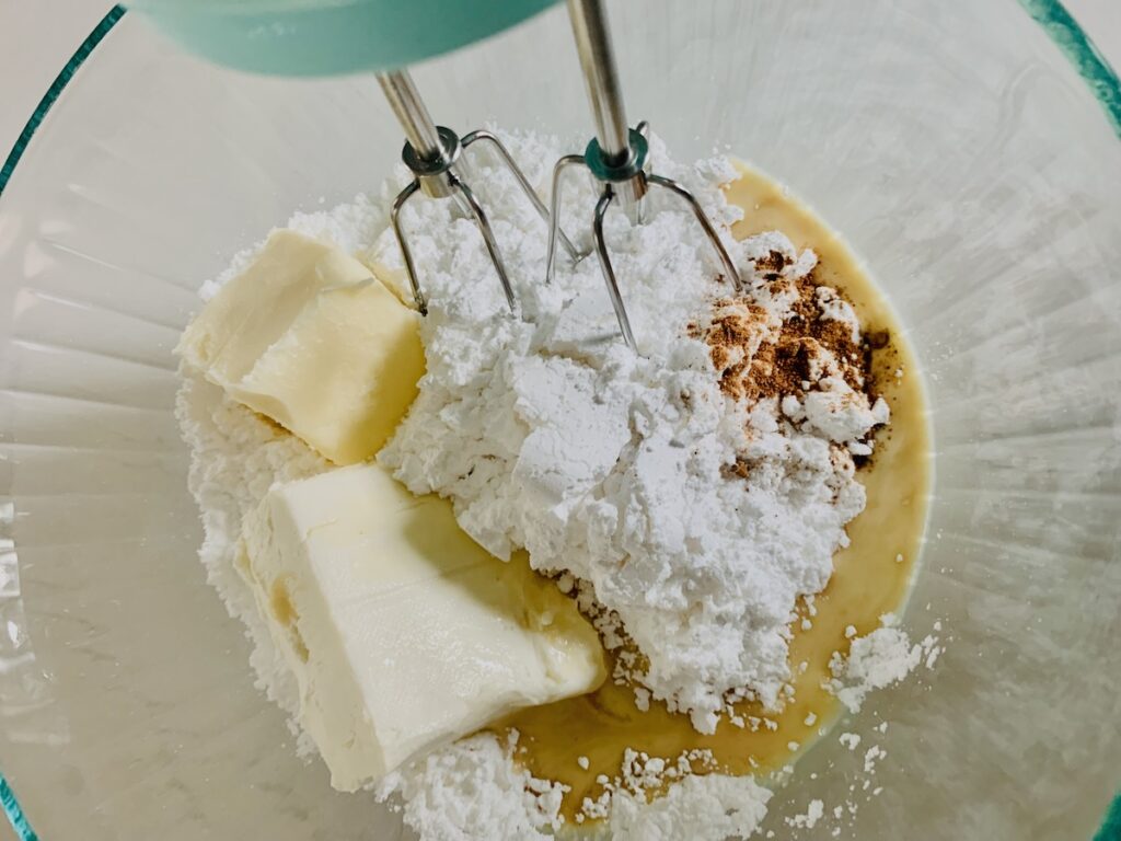 An aqua hand mixer with beaters resting in a bowl of ingredients: powdered sugar, cream cheese, butter, eggnog, vanilla, and nutmeg.