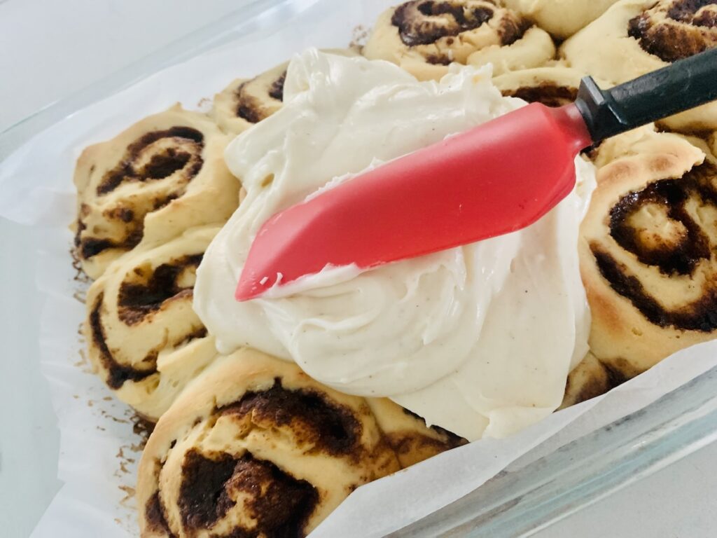 A red spatula spreading eggnog icing on baked gluten-free cinnamon rolls.