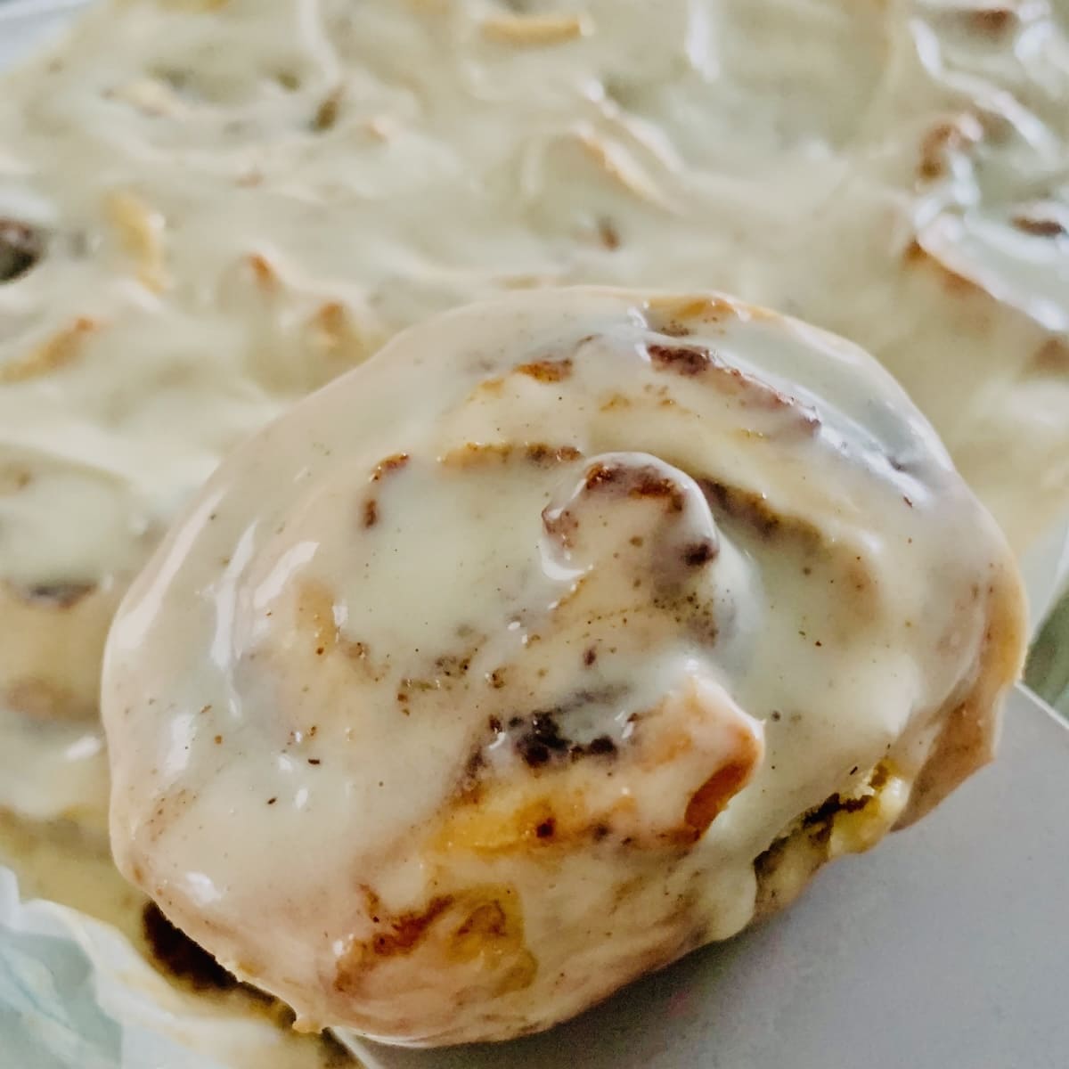 A gluten-free eggnog cinnamon roll held by a grey spatula, with a baking dish of rolls in the background.