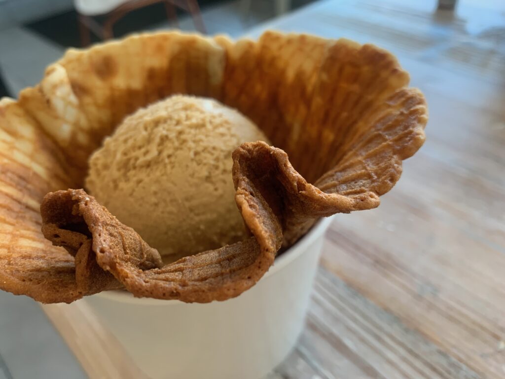 Scoop of caramel-colored ice cream in a gluten-Free waffle cone in a white cup.