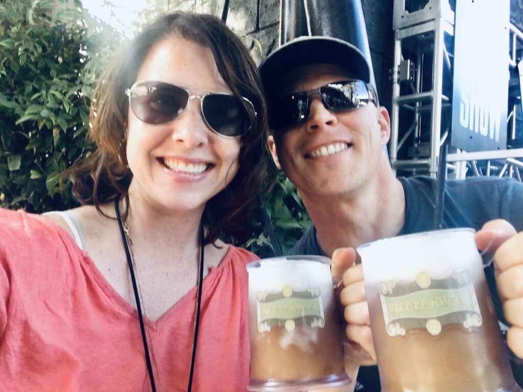 Heather in a bright salmon colored shirt and sunglasses & Dave in a hat and sunglasses holding mugs of gluten-free Butterbeer in souvenir mugs.