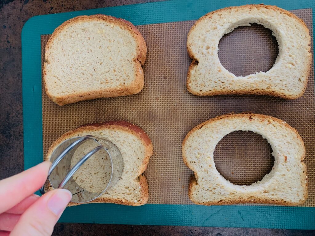 Birds Eye view: four slices of bread on a silicone baking mat on top of a baking sheet. Two have a hole cut out, and one is being cut with a biscuit cutter.