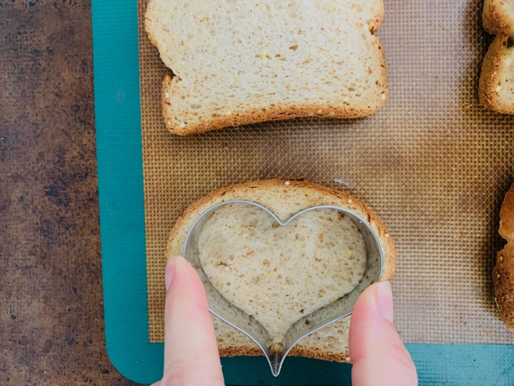 Birds Eye view: slice of bread on a silicone baking mat on top of a baking sheet. Fingers holding a heart-shaped cookie cutter to cut a heart shape out of the side of bread.