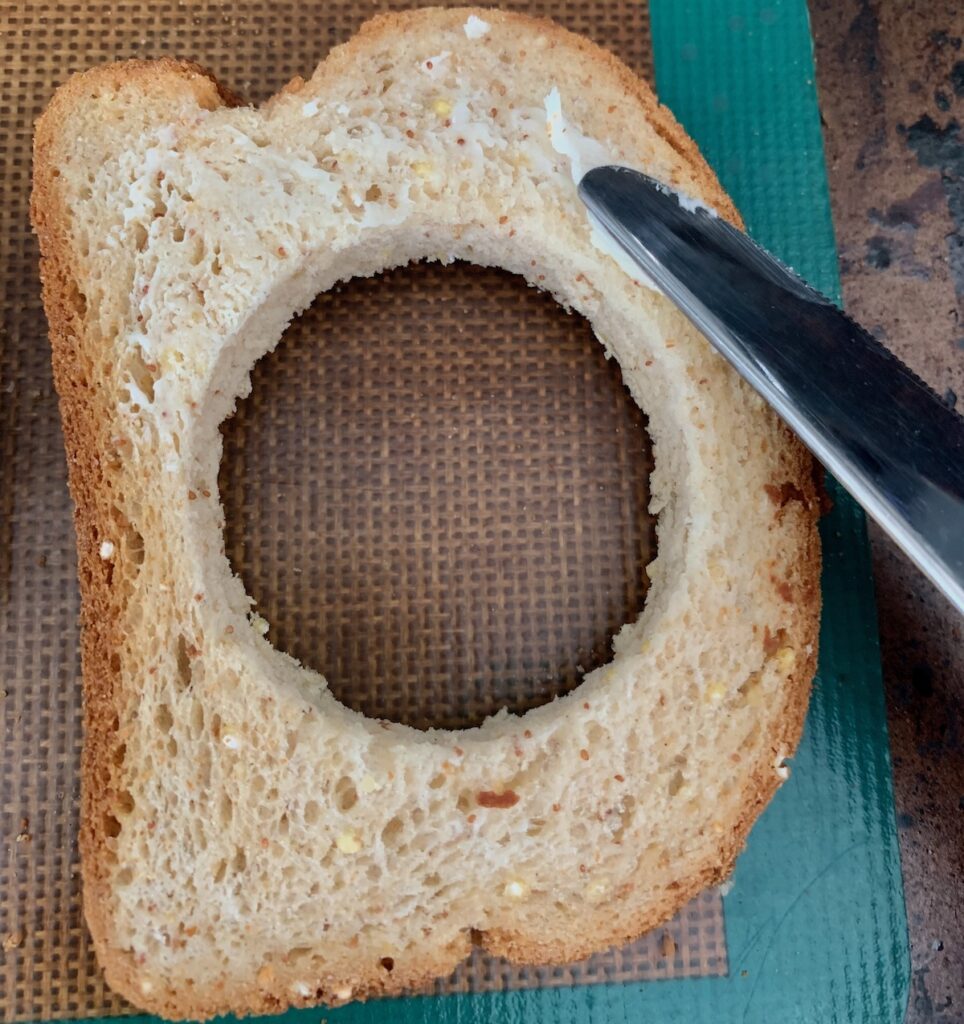 Birds Eye view: knife spreading butter on a slice of bread with a hole cut out of the center.