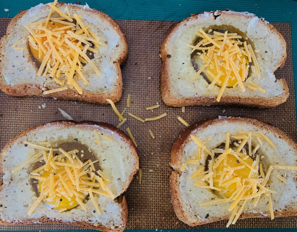 Birds Eye view: four slices of bread with holes cut out and raw egg and shredded cheese on top, lying on a silicone baking mat on top of a baking sheet.