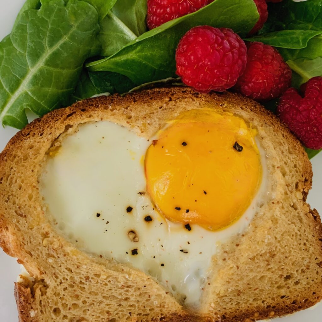 1 slice of gluten-free heart-shaped-egg-in-hole toast topped with cheddar cheese. Spinach and raspberries in the background.