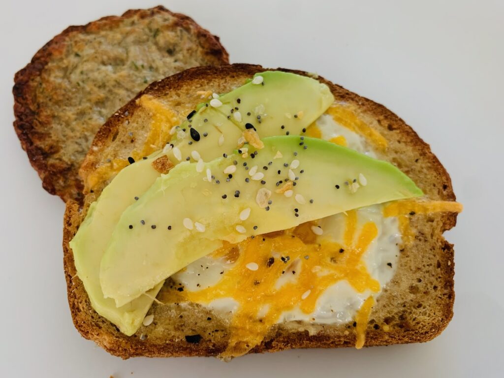 1 slice of gluten-free egg-in-hole toast topped with cheddar cheese, avocado slices and Everything Bagel Seasoning. Sausage patty in the background.