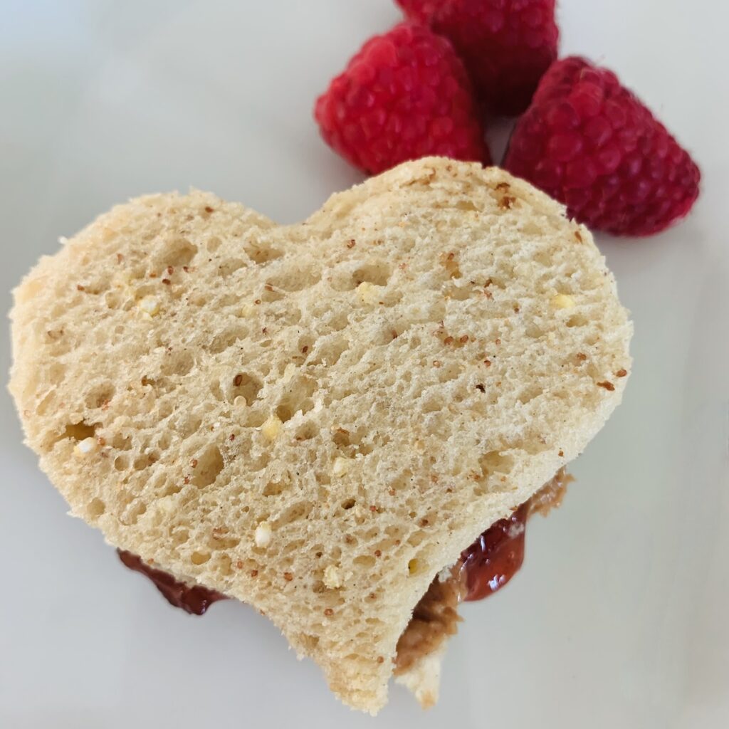 Birds Eye view: heart shaped peanut butter & jelly sandwich, with PB&J dripping out the sides a bit, and 3 raspberries in the background.