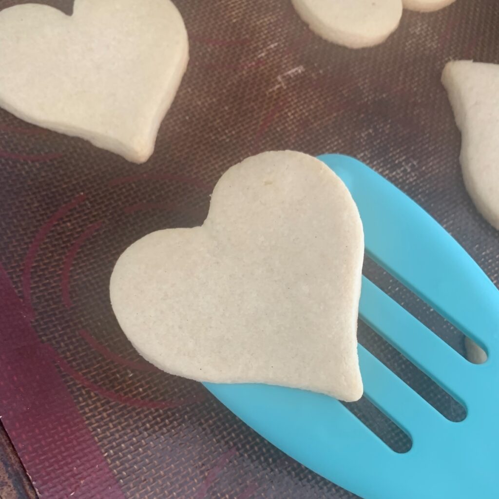 Aqua slotted spatula lifting baked heart cookie off of a silicone baking mat, with a few cookies in the background.