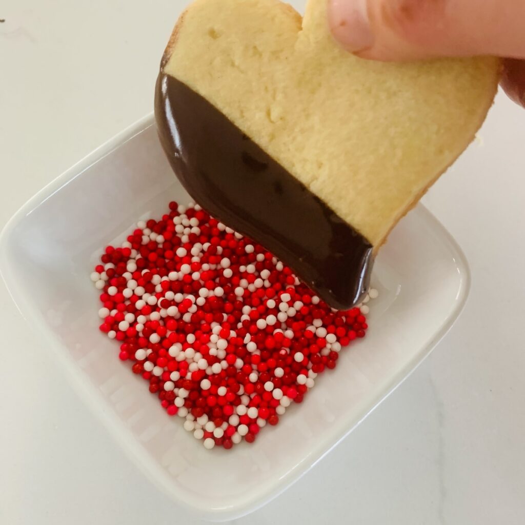 Chocolate dipped heart cookie being dipped in a bowl of red, white, and pink sprinkles.