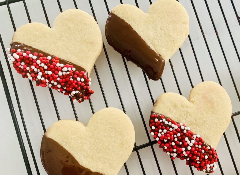 Birds Eye view: four heart cookies on a wire rack. Two dipped in chocolate and 2 dipped in chocolate and sprinkles.