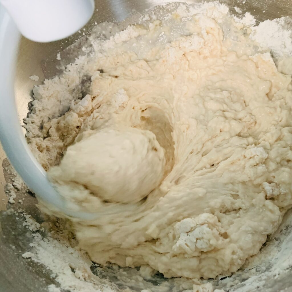 White dough hook mixing dough in a metal mixing bowl.