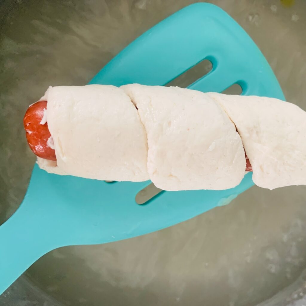 Pretzel wrapped in dough on a teal spatula above boiling baking soda water.