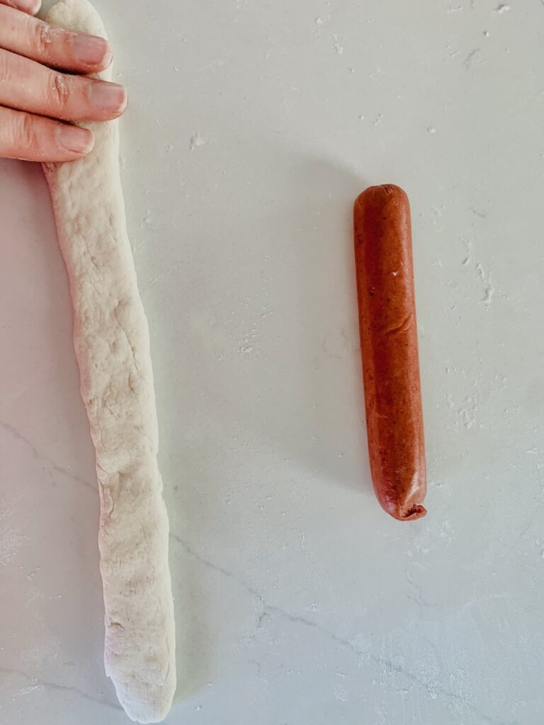 Birds Eye view: patting flat a dough rope next to a hot dog, half the rope's length.