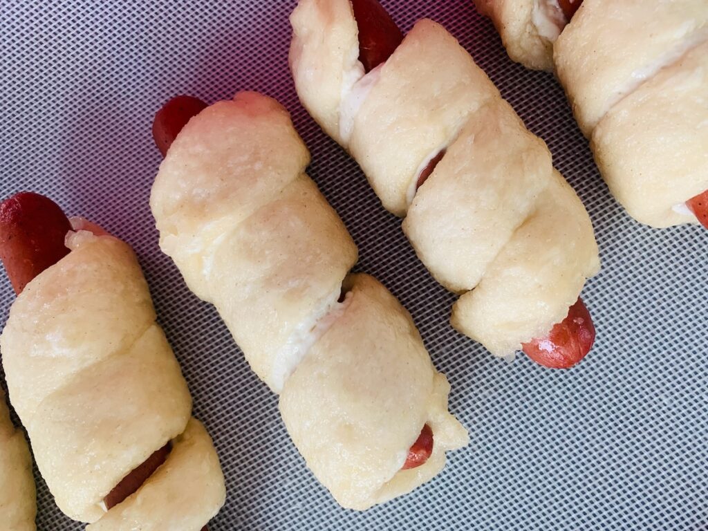 Birds Eye view: hot dogs wrapped in dough (wet and slightly tinted from a baking soda bath) on a silicone baking mat.