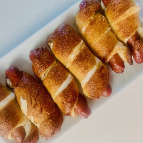 Birds Eye view: a rectangle plate with 6 baked gluten-free pretzel hot dogs.