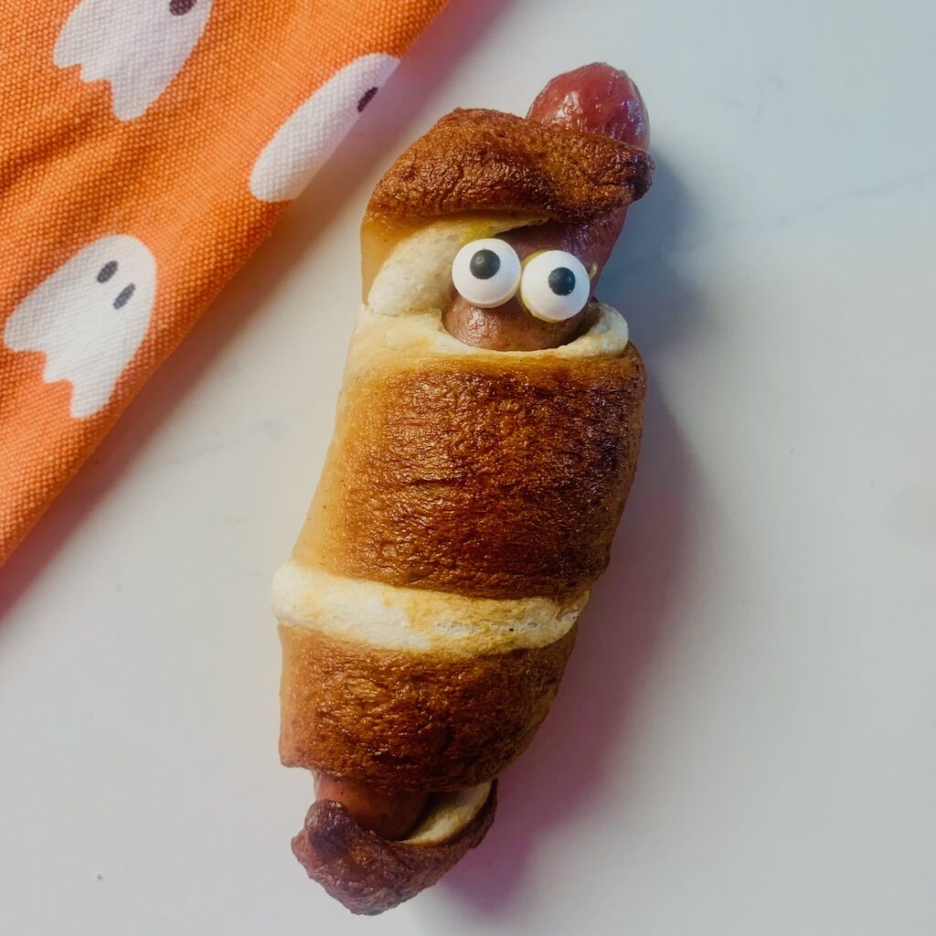 Gluten-free mummy dog wrapped in pretzel bread with candy eyes. An orange dish towel with white ghosts in the background.