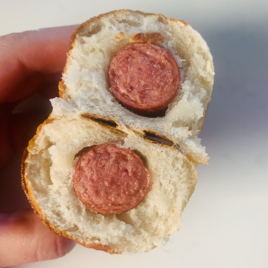 Hand holding a gluten-free pretzel dog cut in off, with a view of the inside of both halves showing a soft bready interior and chewy pretzel exterior.